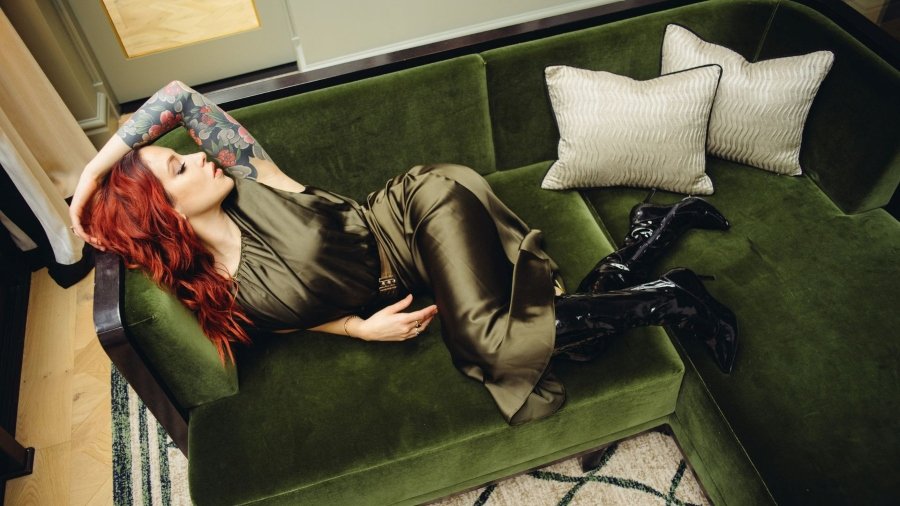 Red-haired woman with tattooed arm reclining on green velvet sofa wearing black boots and satin dress.