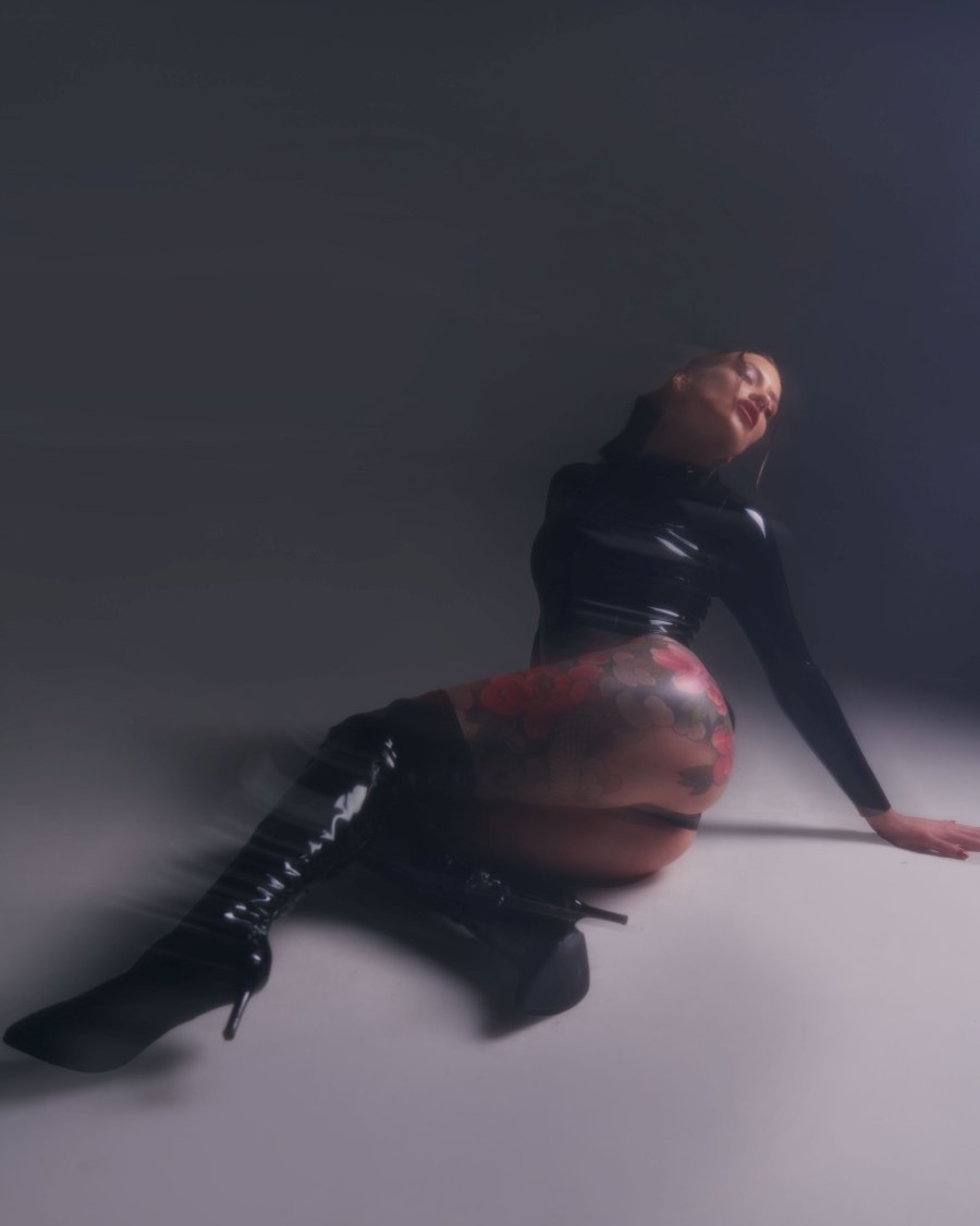 Woman in black latex outfit and thigh-high boots sitting on the floor with a floral tattoo on her thigh.