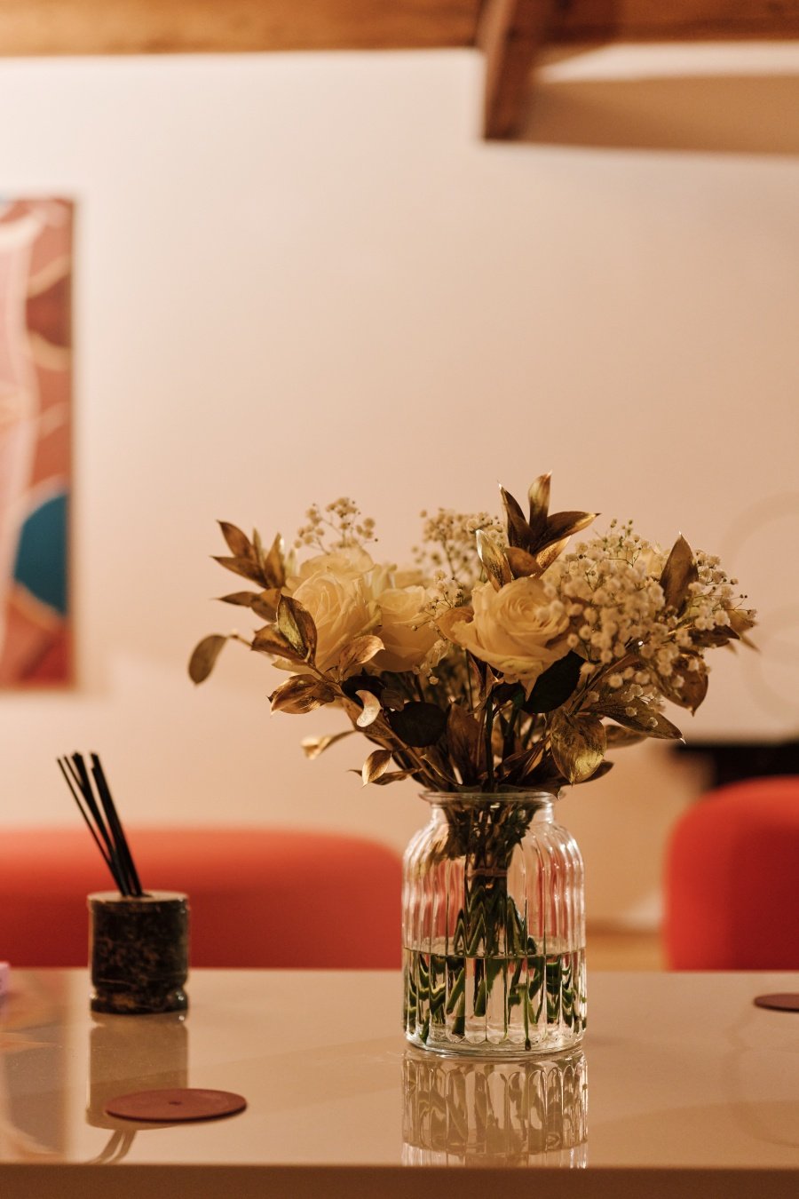 Glass vase with cream roses and gold-tinted leaves on a white table with blurred chairs.