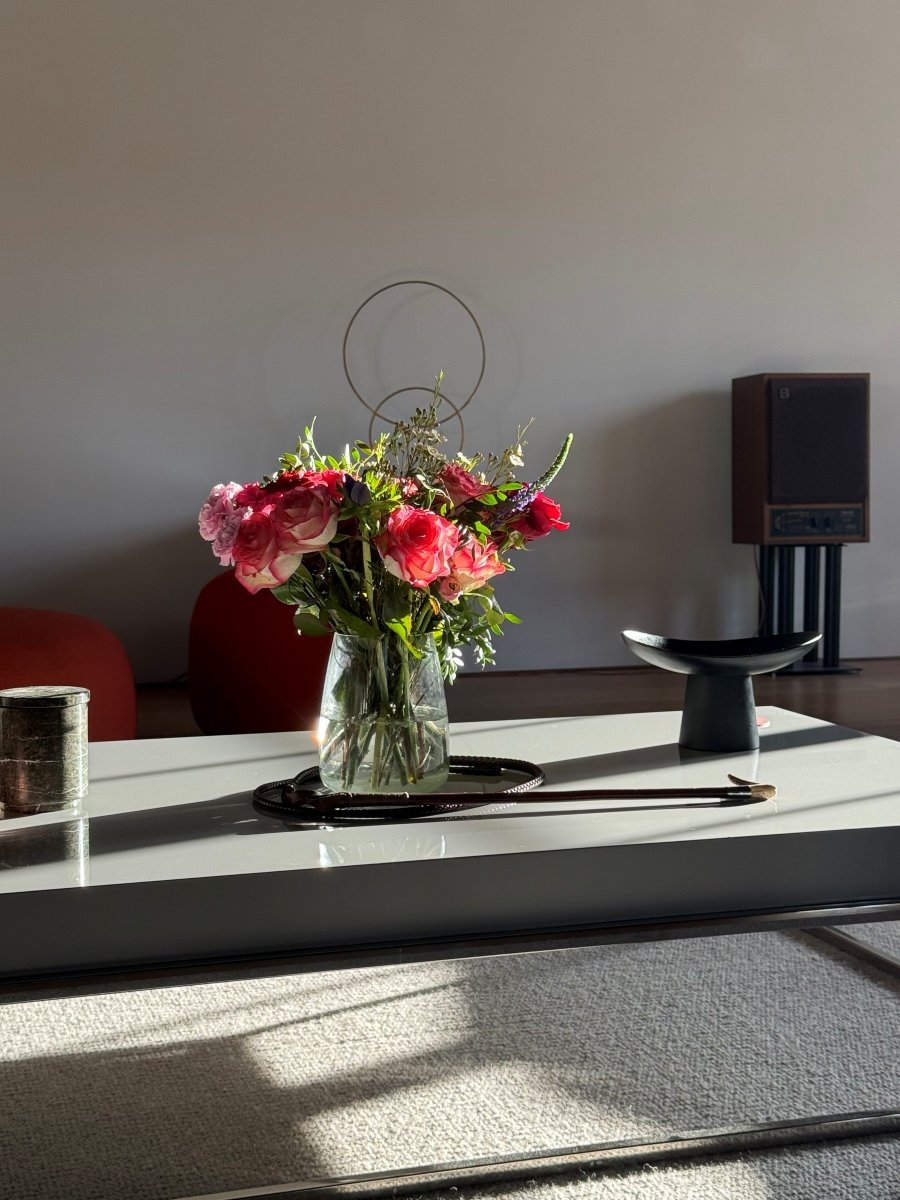 Glass vase with pink and red flowers on white table in modern room lit by sunlight.
